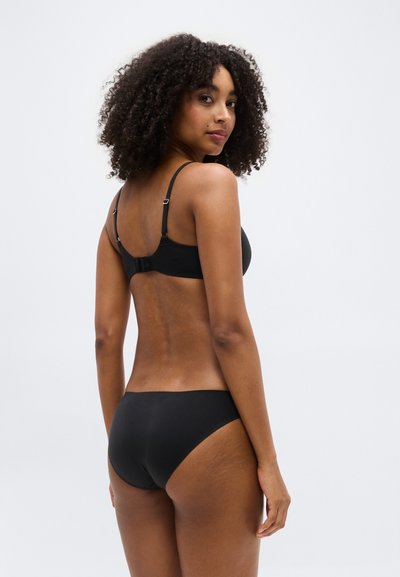 Γυναίκα με κατσαρά μαλλιά που φοράει μαύρο σουτιέν και ταιριαστό εσώρουχο, στέκεται με την πλάτη στην κάμερα, κοιτάζοντας πάνω από τον ώμο της.