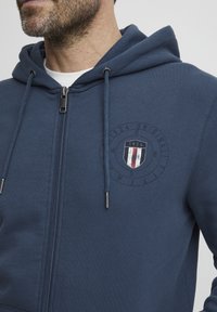 Haut de survêtement zippé bleu marine en tissu doux, doté d'un logo rond avec des accents rouge, blanc et bleu. Comprend des cordons de serrage et une fermeture éclair argentée.