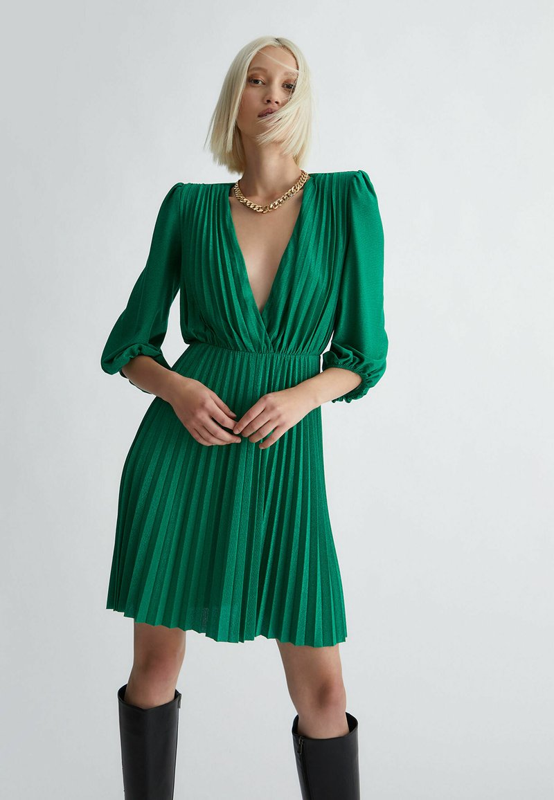 Grün plissiertes Kleid mit tiefem V-Ausschnitt und voluminösen Ärmeln, mit einem gerafften Taillenteil und strukturierter Stoffqualität. Kombiniert mit hohen schwarzen Stiefeln.
