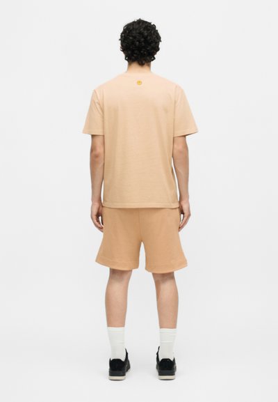 Άνδρας με σγουρά μαλλιά που φοράει μπεζ μπλουζάκι και ταιριαστό σορτς, λευκές κάλτσες και μαύρα αθλητικά παπούτσια, στέκεται με την πλάτη προς τα εμπρός σε λευκό φόντο.