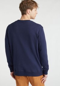 Felpa blu navy con collo rotondo, maniche lunghe e polsini a coste. Tessuto morbido con vestibilità rilassata, abbinata a pantaloni marrone chiaro.
