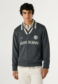 Temno siva pulover s V-izrezom, ki ima bele črte na ovratniku in napis "PEPE JEANS". Prelito čez karirasto srajco. Mehak texture.