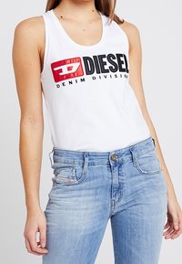 Débardeur blanc avec le logo "DIESEL" en noir et rouge, associé à un jean en denim bleu clair affichant un motif décoloré subtil et des coutures visibles.
