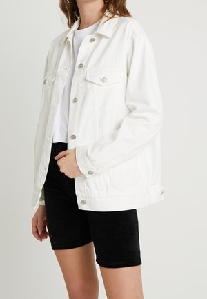 Osoba ubrana w białą, zapinaną na guziki kurtkę jeansową, biały top i czarne krótkie spodenki do kolan na jednolitym tle.