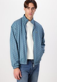 Light blue zip-up jacket featuring a geometric pattern, elastic cuffs, and a small logo detail. Worn over a white crewneck sweater.