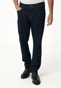 Mörkblå denimjeans med fem fickor, synlig sömnad, knappstängning i midjan och rak bendesign. Bärs med svarta läderstövlar.