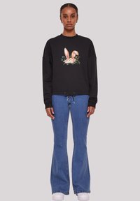 Schwarzes Sweatshirt mit einem grafischen Blumenkaninchen, kombiniert mit blauen Schlagjeans und weißen Schuhen. Der Stoff wirkt weich und lässig.