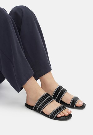Czarne klapki z trzema teksturowanymi paskami ozdobionymi srebrnymi akcentami. Okrągły, otwarty przód, płaska podeszwa, zestawione z szerokimi granatowymi spodniami.