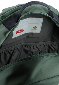 Žalias audinys kuprinės vidus rodo pilką pamušalą, etiketę su Fjällräven logotipu ir elastingą kišenę organizavimui.
