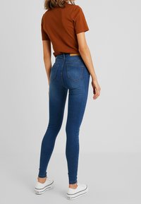 Jeans ajustados de cintura alta en denim azul oscuro, con una textura suave, cinco bolsillos, y combinados con zapatillas blancas.