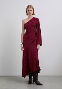 Robe en satin bordeaux à une épaule avec un design drapé, dotée d'une manche longue et d'une fente latérale. Stylisée avec des bottes noires montantes au genou.