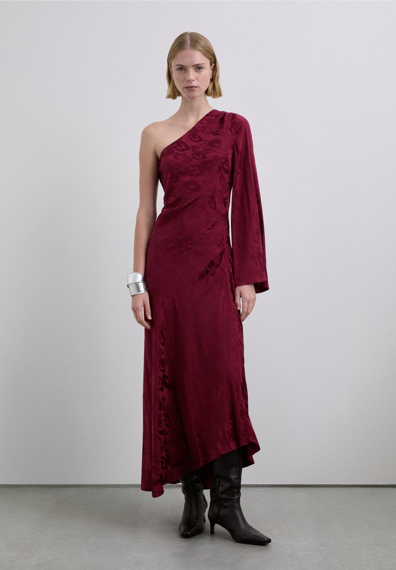 Robe en satin bordeaux à une épaule avec un design drapé, dotée d'une manche longue et d'une fente latérale. Stylisée avec des bottes noires montantes au genou.