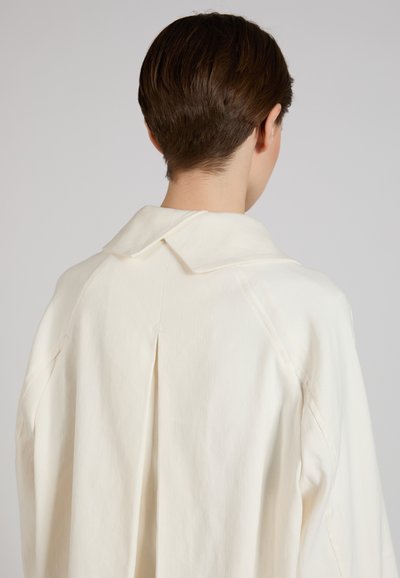 Persona con capelli corti castani vista da dietro che indossa una camicia larga color avorio con un ampio colletto e un plissé sulla schiena.