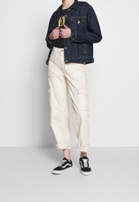 Chaqueta de mezclilla en azul oscuro, acompañada de una camiseta gráfica negra, pantalones cargo crema con bolsillos y zapatillas negras con acentos blancos.