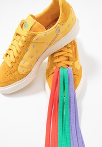 Gula mocka och tyg sneakers med lila broderade detaljer. Inkluderar flerfärgade skosnören: röda, gröna och lila. Platt gummisula.