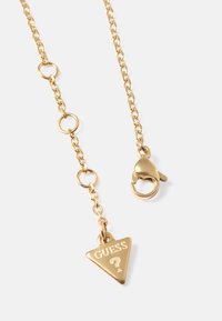 Colar de corrente em ouro com elos ovais, apresentando um pingente triangular gravado com "GUESS" e um ponto de interrogação, acabado com um fecho tipo lagosta.