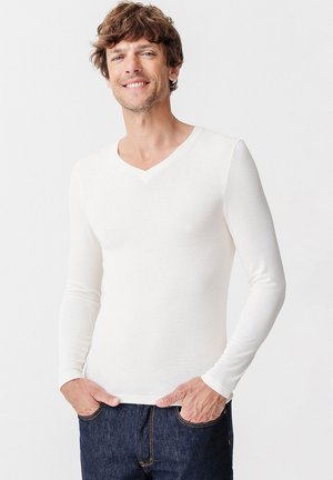 Chemise blanche à manches longues en V, fabriquée dans un tissu doux, au design ajusté, avec une texture lisse. Portée avec un jean en denim bleu.