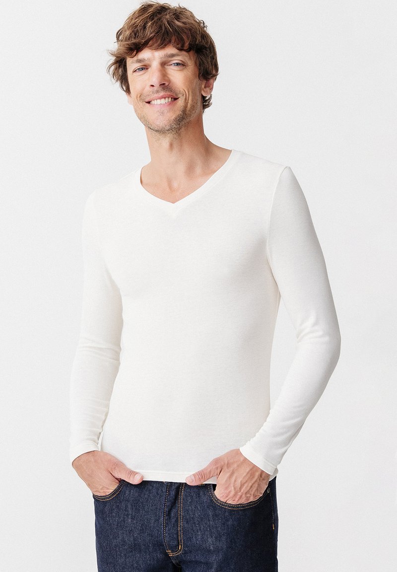Camicia bianca a maniche lunghe con collo a V, realizzata in tessuto morbido, design aderente e texture liscia. Indossata con jeans in denim blu.