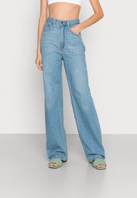 Jeans de ganga de cintura alta e perna larga, de cor azul clara, com bolsos frontais e fecho com um único botão, combinados com sandálias de trama verde e de calçar facilmente.