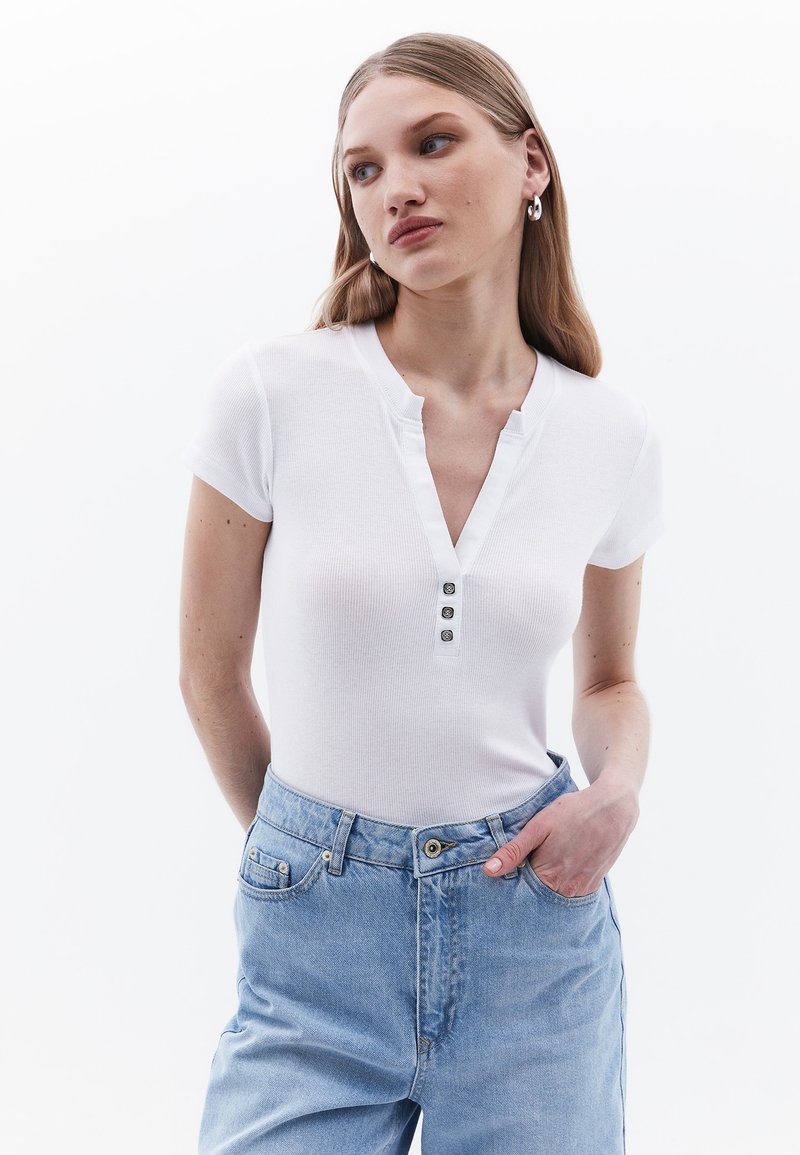 Top corto a maniche corte bianco a coste con scollatura a V e chiusura a tre bottoni. Abbinato a jeans a vita alta azzurri, in evidenza una vestibilità comoda.