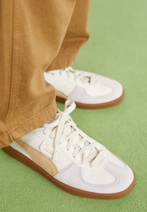 Białe i beżowe buty sportowe Puma z gumową podeszwą, noszone z brązowymi spodniami na zielonym, wykładanym dywanie.