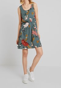 Květinové A-čkové šaty v teal barvě se vzory v růžové, červené a oranžové. Bez rukávů, s přiléhavým pasem a splývavou sukní, doplněné bílými teniskami.