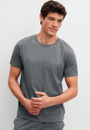 Mann mit kurzen dunklen Haaren trägt ein schlichtes graues Kurzarm-T-Shirt und steht vor einem weißen Hintergrund mit verschränkten Händen.