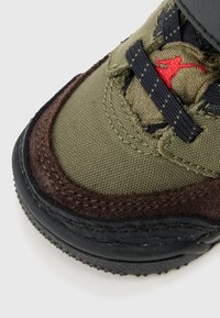 Groene en bruine sneaker met een gestructureerde bovenkant, contrasterende stitching, een zwarte rubberen zool en rode logo-accenten op de veters.