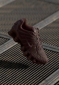 Tamno smeđi Nike teniski/tenisice s teksturiranim mrežastim gornjištem, prepoznatljivim valovitim dizajnom, podstavljenim potplatom i čipkastim prednjim dijelom, prikazani na mrežastoj površini.