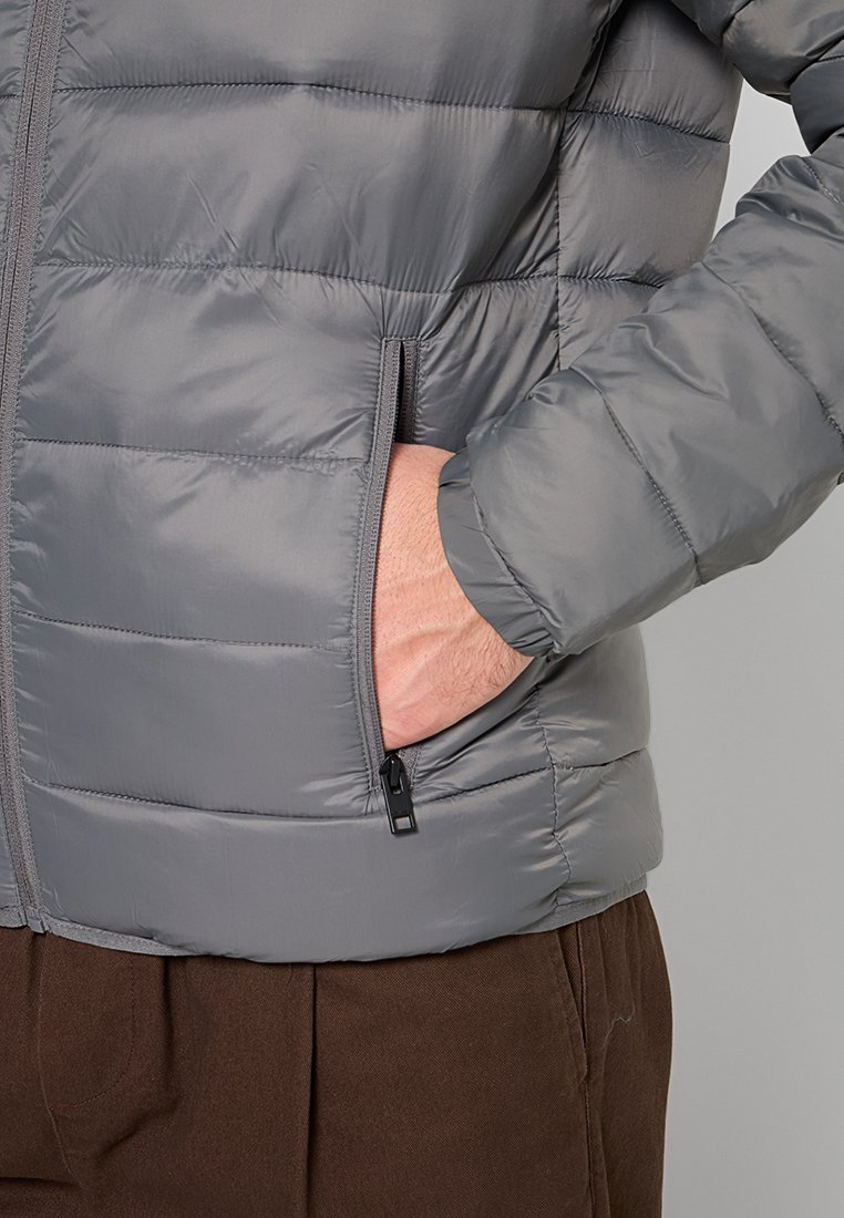 Chaqueta tipo puffer gris con textura acolchada, que cuenta con un bolsillo con cremallera y un cuello alto. Puños y dobladillo elásticos para un mejor ajuste.