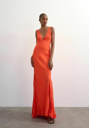 Femme portant une robe orange sans manches, longue jusqu'au sol, avec un décolleté en V profond et des détails en dentelle, debout contre un mur blanc uni.