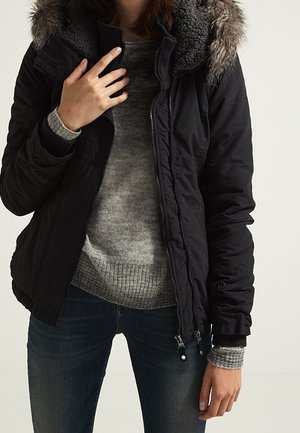 Personne portant une veste d’hiver noire avec capuche bordée de fourrure sur un pull gris en maille et un jean bleu, ajustant le col de la veste.