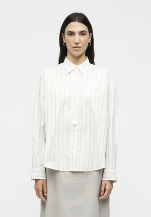 Γυναίκα με μακριά σκούρα μαλλιά που φορά λευκό πουκάμισο με λεπτές ρίγες και ανοιχτό γκρι φούστα, στέκεται μπροστά σε απλό φόντο.