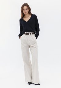 Jersey de pico en navy combinado con pantalones de pierna ancha beige; cinturón marrón con hebilla de plata; el modelo está de pie con las manos en los bolsillos.