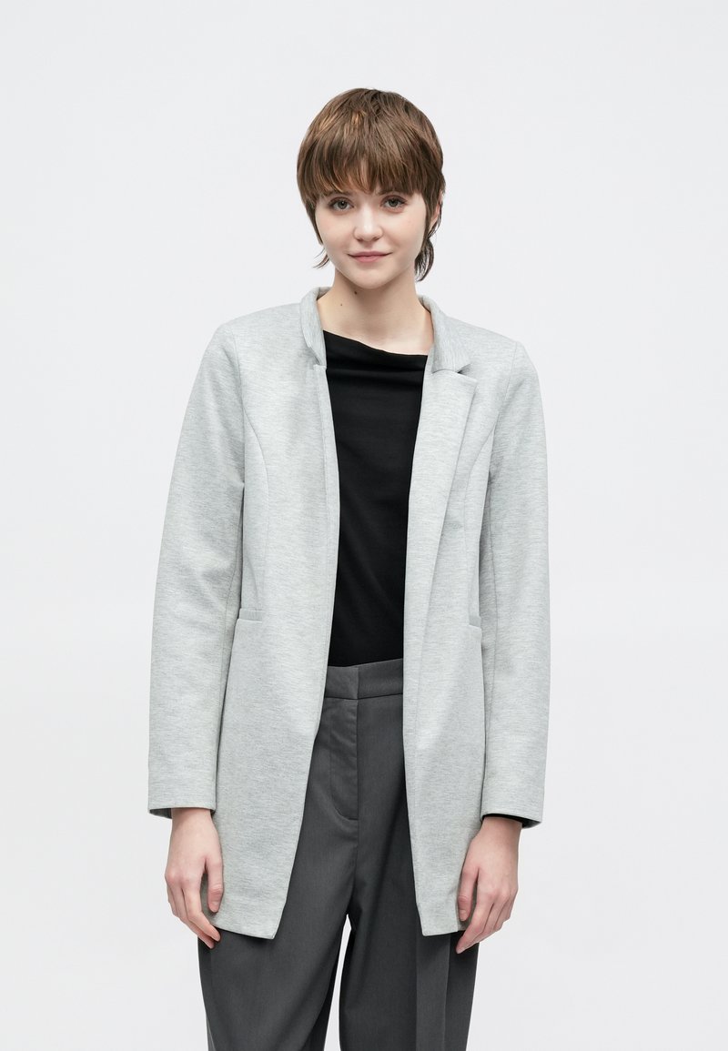 Szürke, hosszú ujjú blézer puha anyagból, laza szabással, felhajtott gallérral és két elülső zsebbel, fekete felsővel és szürke nadrággal párosítva.