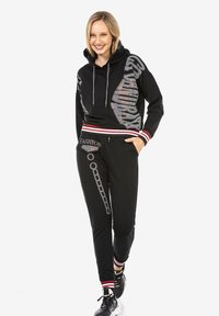 Schwarzer Sweatshirt mit Strassverzierungen, mit gestreiftem Saum und Bündchen. Kombiniert mit schwarzen Hosen und sportlichen Schuhen.