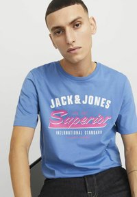 Blå bomulds t-shirt med korte ærmer og rund halsudskæring. Har hvid og pink grafisk tekst "JACK & JONES Superior International Standard."