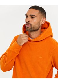 Sudadera con capucha de forro polar naranja con bolsillo tipo canguro y puños acanalados; presenta un pequeño logo bordado en el pecho; textura suave.