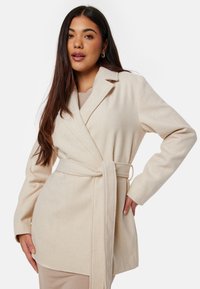 Cappotto avvolgente beige con maniche lunghe, colletto a revers e vita legata. Realizzato in un materiale morbido e strutturato. Presenta tasche laterali.