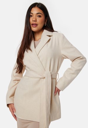 Manteau enveloppant beige avec des manches longues, un col à revers et une taille nouée. Fabriqué dans un matériau doux et texturé. Possède des poches latérales.