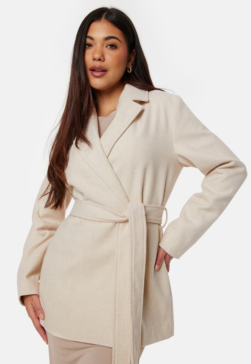 Cappotto avvolgente beige con maniche lunghe, colletto a revers e vita legata. Realizzato in un materiale morbido e strutturato. Presenta tasche laterali.