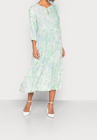 Vestido midi floral en azul claro y verde, con escote en pico con bordes festoneados, mangas tres cuartos y un dobladillo con volantes en capas. Llevado con tacones blancos.