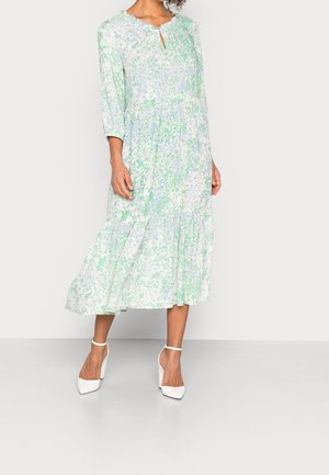Florale midi-jurk in lichtblauw en groen, met een golvende halslijn, driekwart mouwen en een gelaagde rucheszoom. Draagt witte hakken.