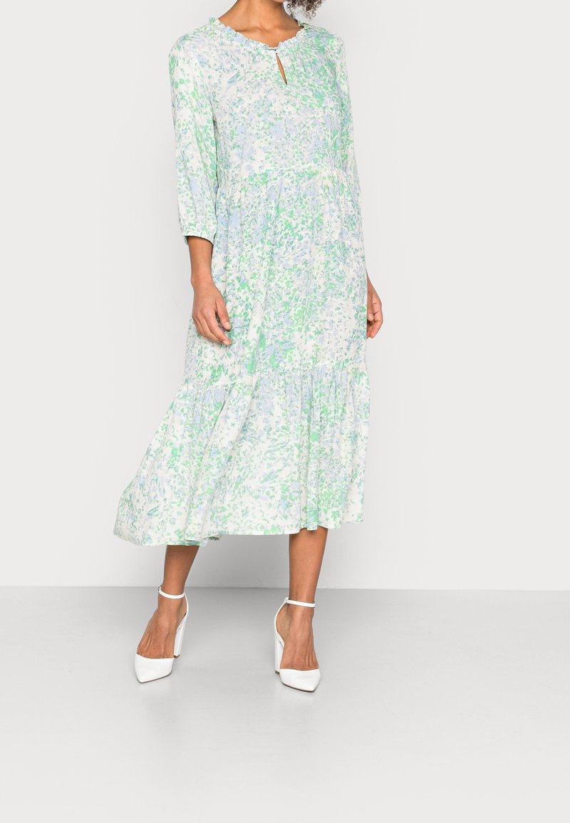Vestido midi floral en azul claro y verde, con escote en pico con bordes festoneados, mangas tres cuartos y un dobladillo con volantes en capas. Llevado con tacones blancos.