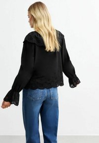 Svart blus med broderad scalloped nederkant och ärmar. Materialet verkar lätt och har en avslappnad passform. Matchas med blå denimjeans.