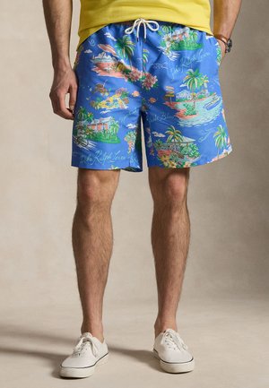 Bărbat purtând pantaloni scurți albaștri cu imprimeu tropical cu palmieri, scene de plajă și șnur alb, asortați cu pantofi sport albi și tricou galben.