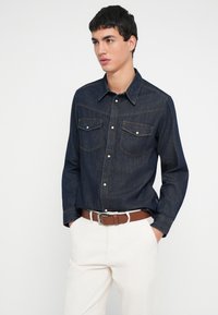 Fiatal férfi sötétkék farmer ingben, mely gombos és mellzsebekkel rendelkezik, fehér nadrág barna övvel, kezét a zsebében tartva, jobbra néz.