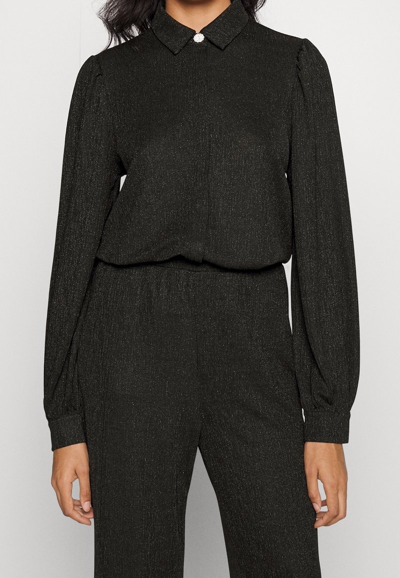 Femme portant un haut noir à manches longues avec un petit col et un pantalon noir assorti en tissu texturé.
