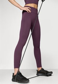 Lila bordás leggings magas derékkal, fekete hálós sportcipővel párosítva. Egy ellenállás-szalag helyezkedik el a lábak között.