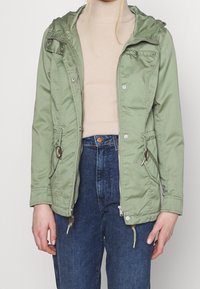 Persona che indossa una giacca leggera verde chiaro con zip e tasche sopra un dolcevita beige e jeans a vita alta blu, in piedi contro uno sfondo semplice.